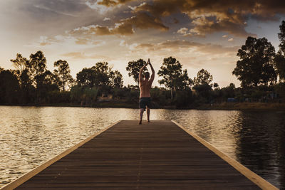 Rear view of man doing yoga on pier over lake at sunset