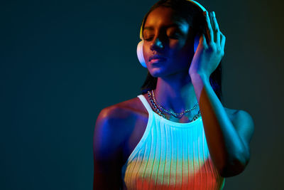 Serious ethnic brazilian female listening to music in modern wireless headphones while standing with eyes closed in studio with fluorescent light