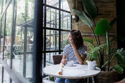 Woman looking through window while talking on mobile phone at cafe