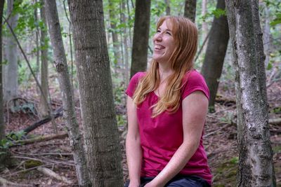 Woman smiling while sitting by tree trunk in forest