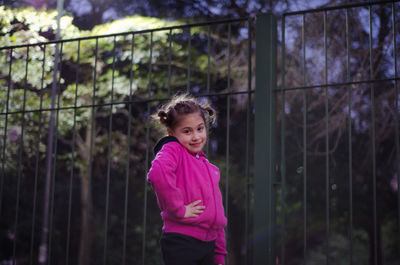 Low angle portrait of girl standing by fence at dusk