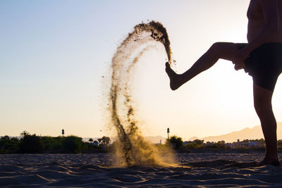 Low section of man kicking sand while standing at shore of beach during sunset