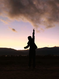 Silhouette man with hand raised standing on field against sky during sunset