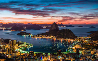 Illuminated city by sea against sky at sunset