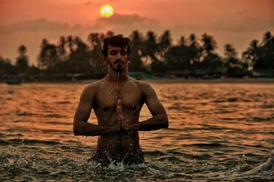 Shirtless man standing in praying position during sunrise on water