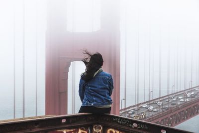 Rear view of woman sitting on railing against bridge during foggy weather