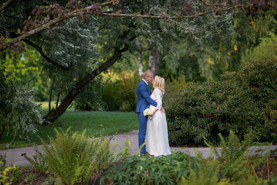 Side view of wedding couple kissing while standing on footpath amidst plants at park