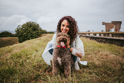 Portriat of woman with dog crouching on grass against sky