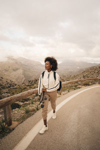 Woman looking away while walking on road holding skateboard against sky