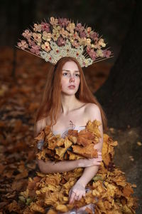 Thoughtful young woman covered with leaves sitting at forest during autumn