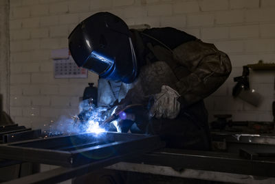 Unrecognizable male employee in protective gloves and helmet using welding machine while working in dark workshop