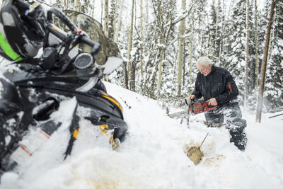 Retired elderly man using chainsaw to clear trails while snowmobiling.