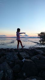 Side view of girl walking on rocky beach against sky during sunset