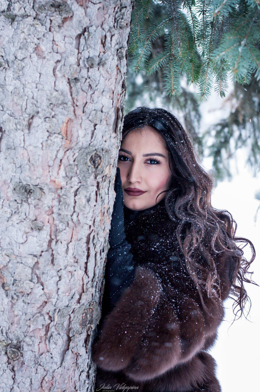 tree trunk, tree, looking at camera, one person, portrait, real people, beautiful woman, leisure activity, young adult, smiling, long hair, young women, nature, day, outdoors, happiness, lifestyles, winter, close-up, people