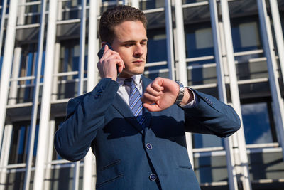 Low angle view of businessman talking on phone while checking time in city