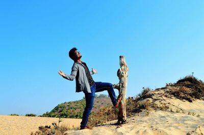 Full length of young man with arms outstretched standing on desert against clear blue sky