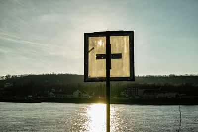 Cross shape road sign by river against sky