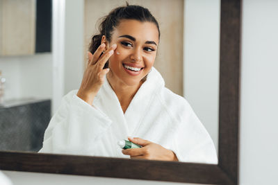 Smiling young woman applying cream on face while looking in mirror