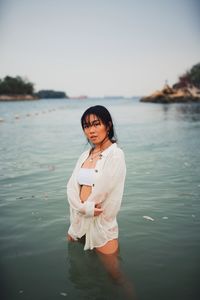 Portrait of young woman wearing bikini standing in sea against clear sky