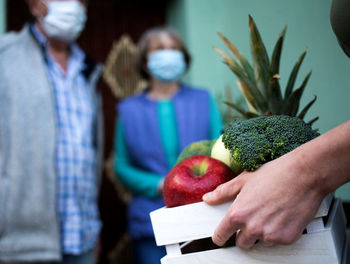 Hand of woman carrying crate with food for senior couple during pandemic