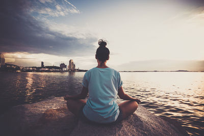 Rear view of woman meditating while sitting on rock in sea