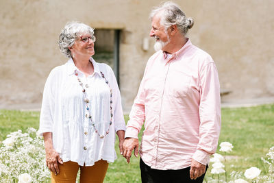 Content elderly couple looking at each other while standing holding hands on street with decoration near building during wedding party on summer day