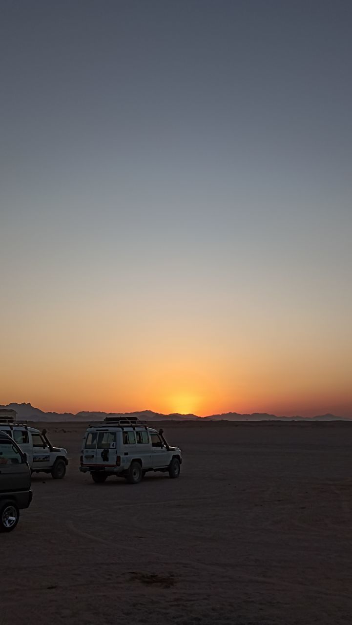 horizon, sky, sunset, mode of transportation, natural environment, transportation, desert, car, motor vehicle, nature, dawn, scenics - nature, land, beauty in nature, land vehicle, landscape, no people, copy space, travel, environment, orange color, sea, tranquility, vehicle, sand, clear sky, tranquil scene, outdoors, non-urban scene, evening, travel destinations, off-road vehicle