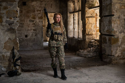 Blonde woman in army uniform holding a firearm in an abandoned building