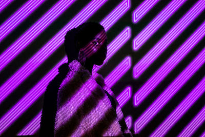 African american woman in stylish fur coat looking at camera while standing under violet neon stripes