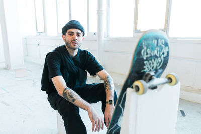 Portrait of young man sitting with skateboard by window