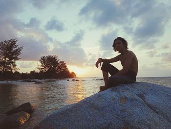 Man sitting on rock at shore against sky during sunset