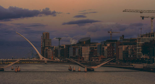 Bridge over river by buildings against sky during sunset