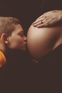 Close-up of boy kissing on pregnant woman abdomen against black background