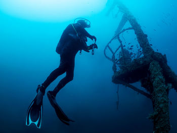 Professional diver with flashlight swimming near old sunken ship covered with moss on bottom of deep sea with clear water