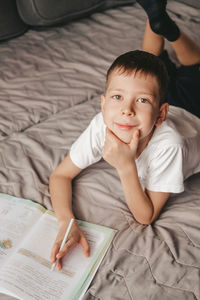 Portrait of boy doing homework lying on gray sofa. a teenager writes in a workbook on the couch.