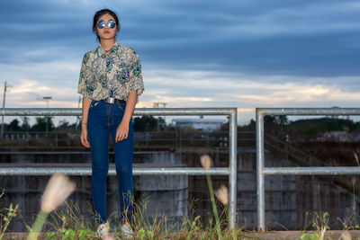 Young woman standing by railing against sky