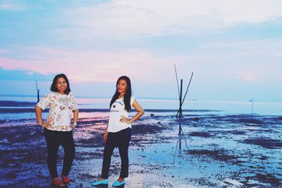 Portrait of female friends standing at beach against sky during sunset