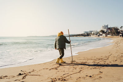 Boy in yellow rubber boots playing with water and sand at the beach. child kid at autumn winter sea.