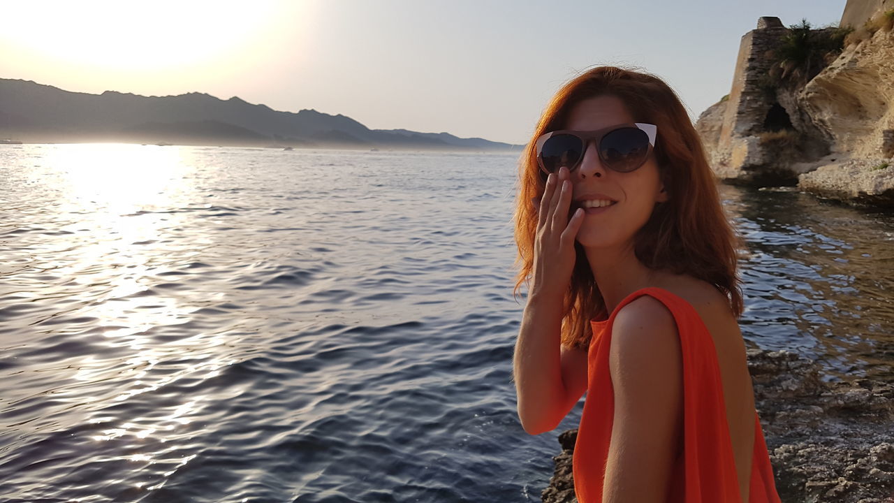 water, sunglasses, real people, sea, one person, nature, young adult, young women, leisure activity, lifestyles, smiling, outdoors, portrait, sunset, looking at camera, beautiful woman, mountain, beauty in nature, beach, happiness, day, sky, clear sky, people