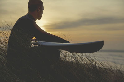 Side view of man sitting on shore against sunset sky