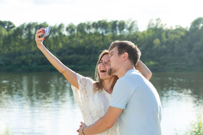 Young couple doing selfie while standing against lake