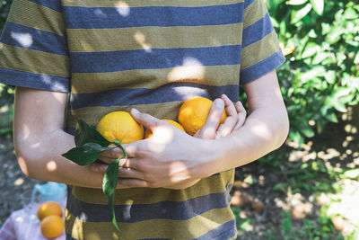 The boy stands among the trees in the garden and holds ripe oranges in his hands.