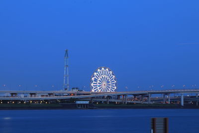 Illuminated ferris wheel by river against clear blue sky