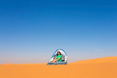 Mature woman sitting in tent on sand at desert against clear blue sky