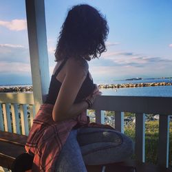 Woman sitting on porch while looking at lake against sky