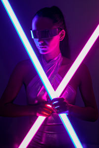 Self assured young female model with long hair in futuristic outfit and vr goggles standing in dark room with neon swords in hands