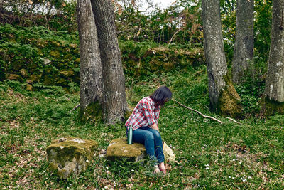 Woman sitting in forest on stone to rest, traveling with backpack in a grove or exercising outdoors