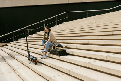 Woman talking on mobile phone while sitting by saxophone instrument and electric push scooter on staircase