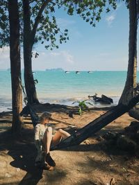 Woman sitting on tree by sea against sky