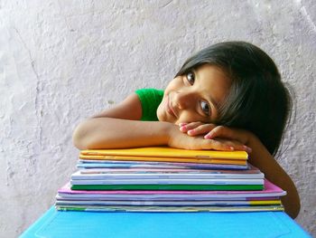 Portrait of smiling girl with stacked books sitting against wall at home